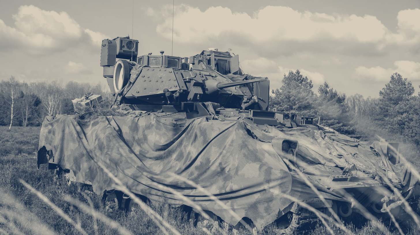 Two-tone U.S. Army photo of an M2A4 Bradley fighting vehicle under camouflage netting in a field in Poland.