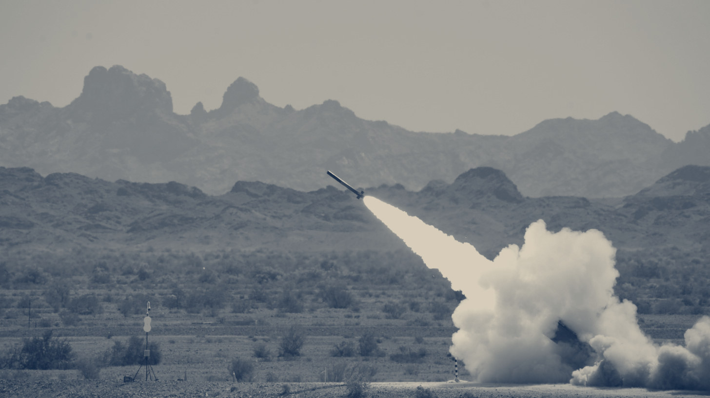 Two-tone U.S. Army photo of the Autonomous Multi-Domain Launcher firing a practice rocket at Yuma Proving Ground, mountains behind.