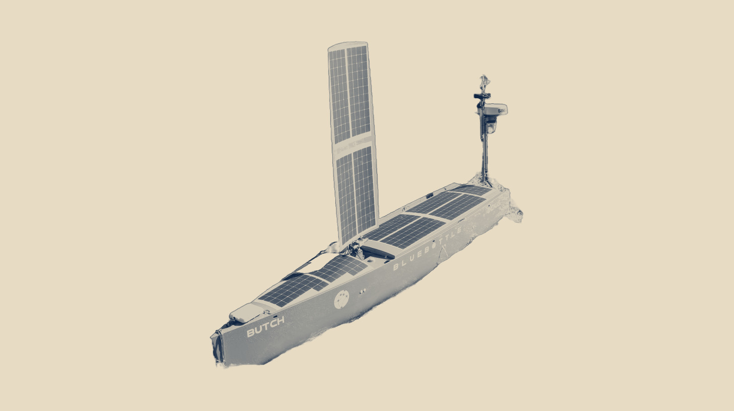 Two-tone photo of an Ocius Bluebottle uncrewed surface vessel under sail, an Australian autonomous maritime program, as a stand-in; it is not the Ghost Shark, on a plain sand background.