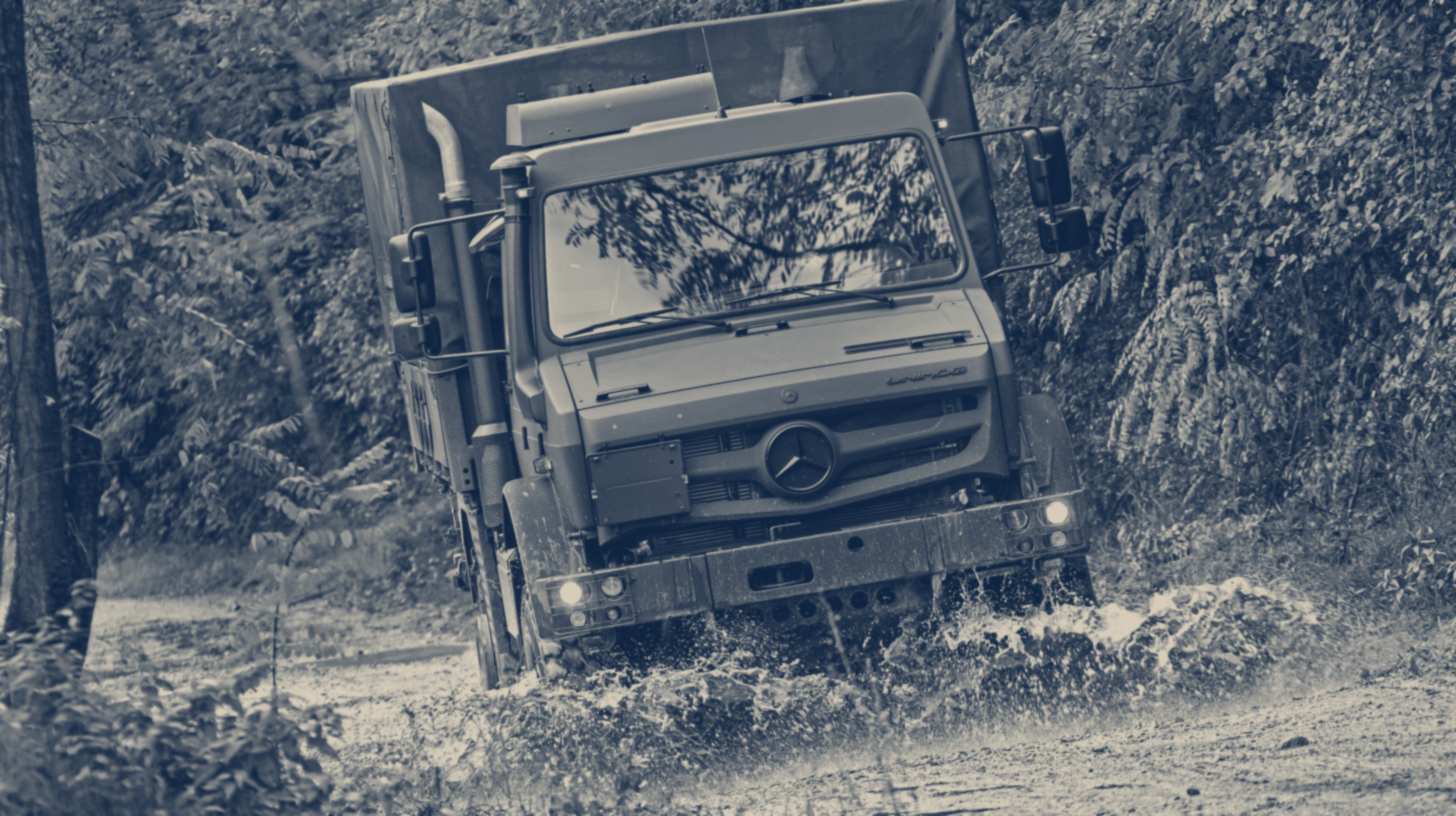 Two-tone Daimler Truck photo of a Mercedes-Benz Unimog driving through mud on a forest track.