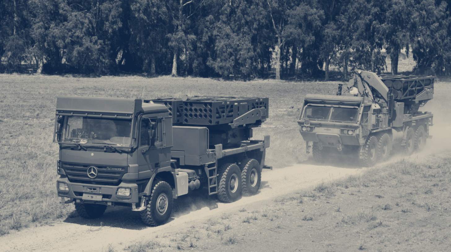 Two-tone Elbit Systems company photo of two PULS rocket launchers on 6x6 trucks driving along a dirt track; the truck variant may differ from the contract configuration.