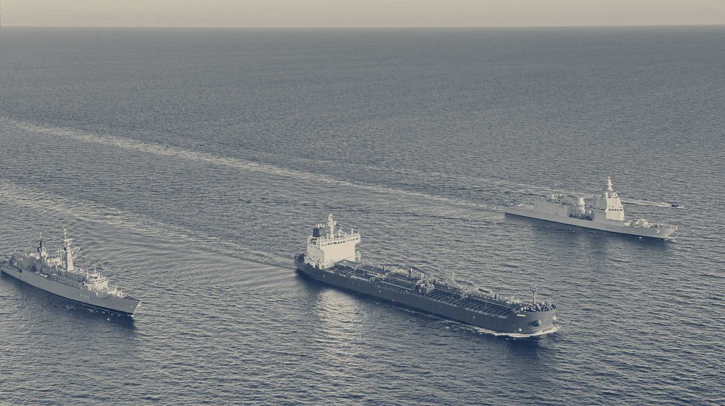 Two-tone EUNAVFOR MED IRINI photo of Operation IRINI warships shadowing a tanker at sea.