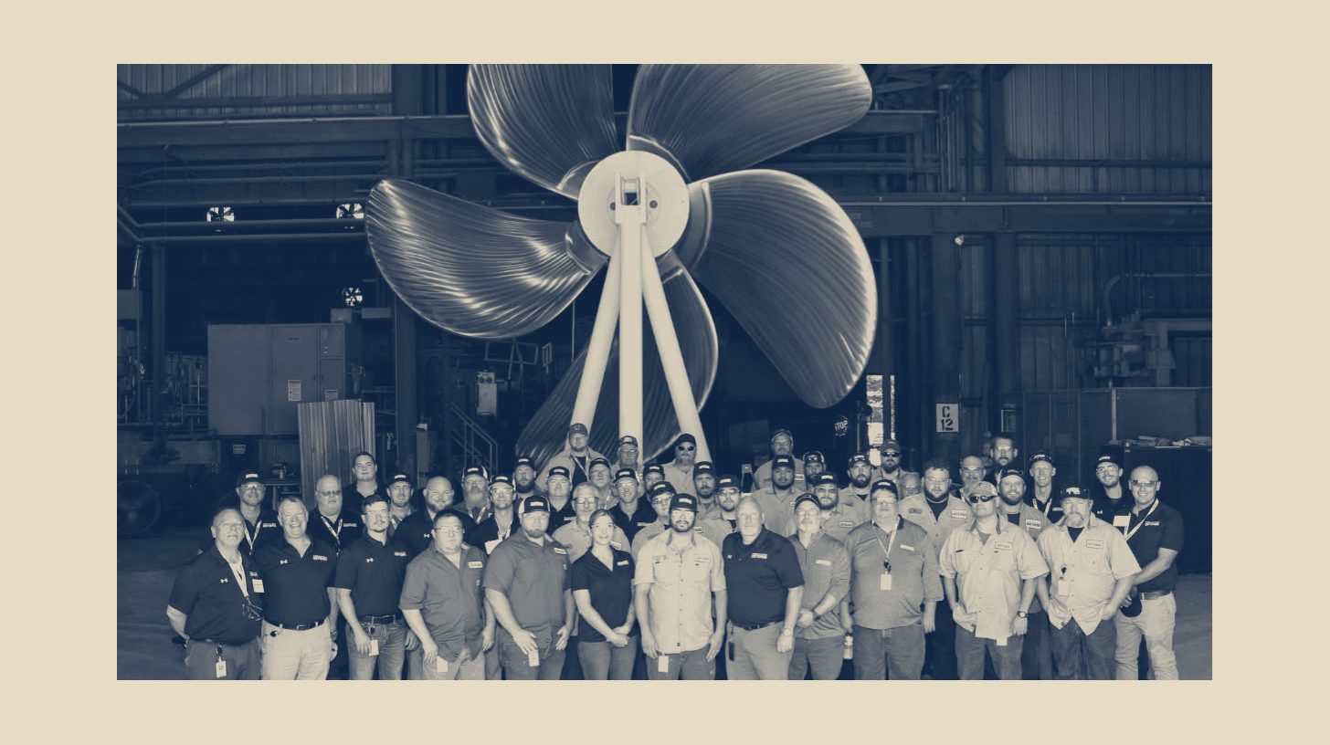 Two-tone Fairbanks Morse Defense company photo of the Pascagoula workforce posed beneath a large naval propeller, shown as a panel on a plain sand background.