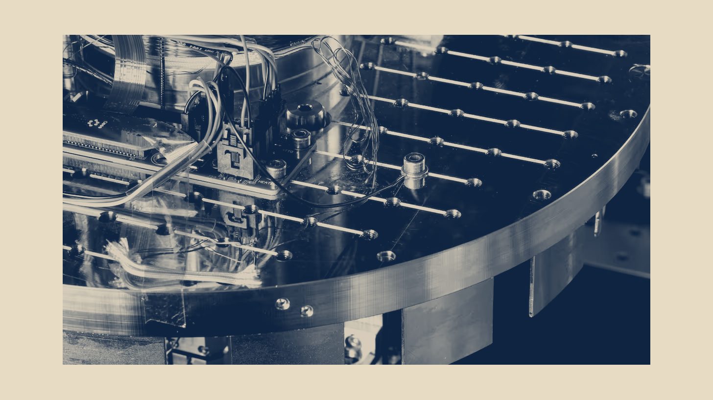 Two-tone IonQ photo of quantum computer hardware, a copper cold-stage plate with wiring and circuit boards, shown as a panel on a sand background.