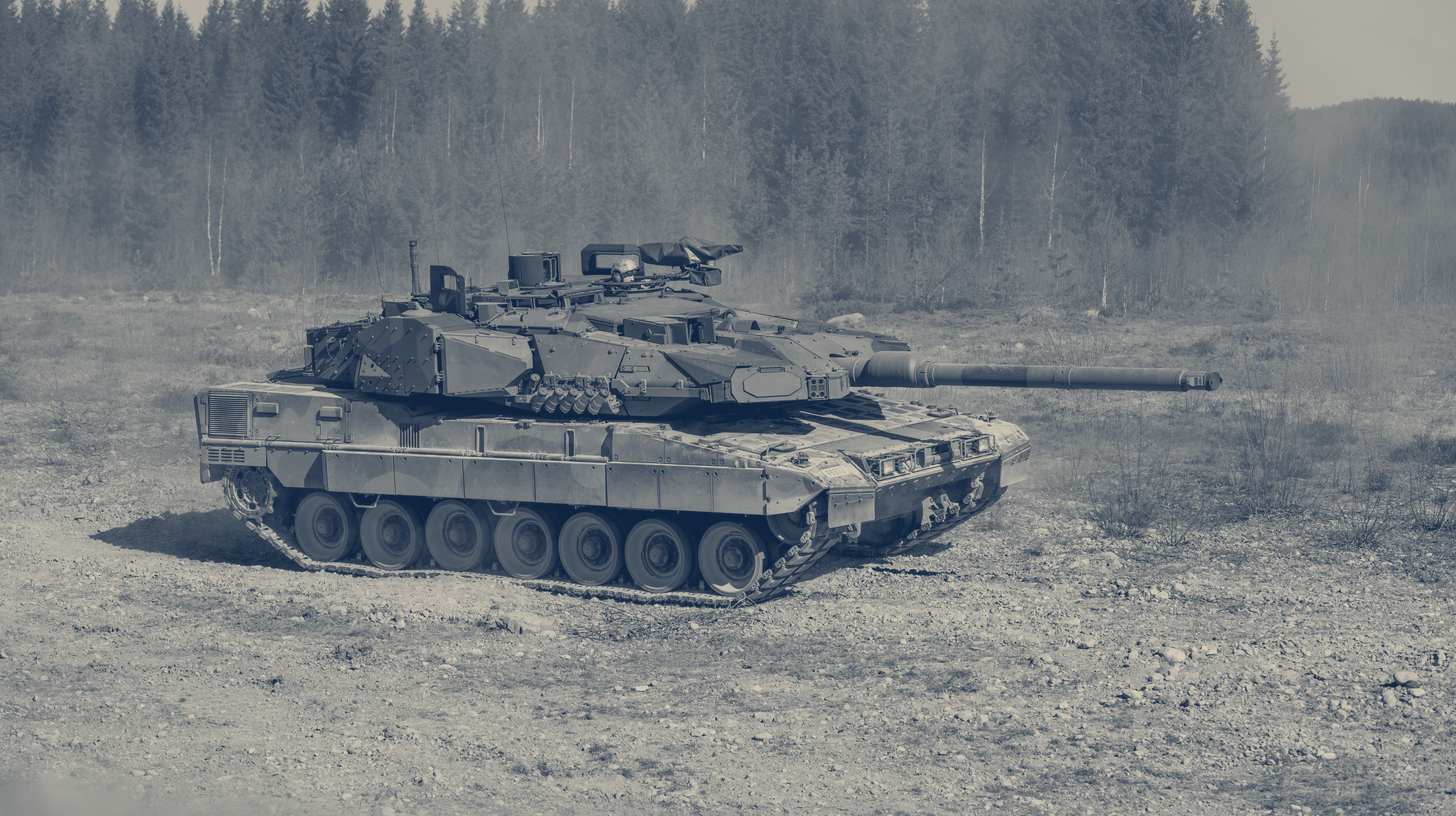 Two-tone photo of a Norwegian Leopard 2A8 NO tank on the move across open ground, trees behind.
