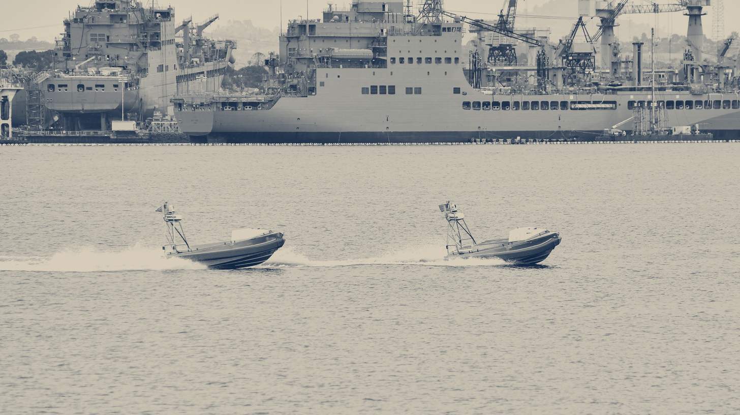 Two-tone U.S. Navy photo of two Global Autonomous Reconnaissance Craft uncrewed boats running at speed off Coronado, Navy ships behind.