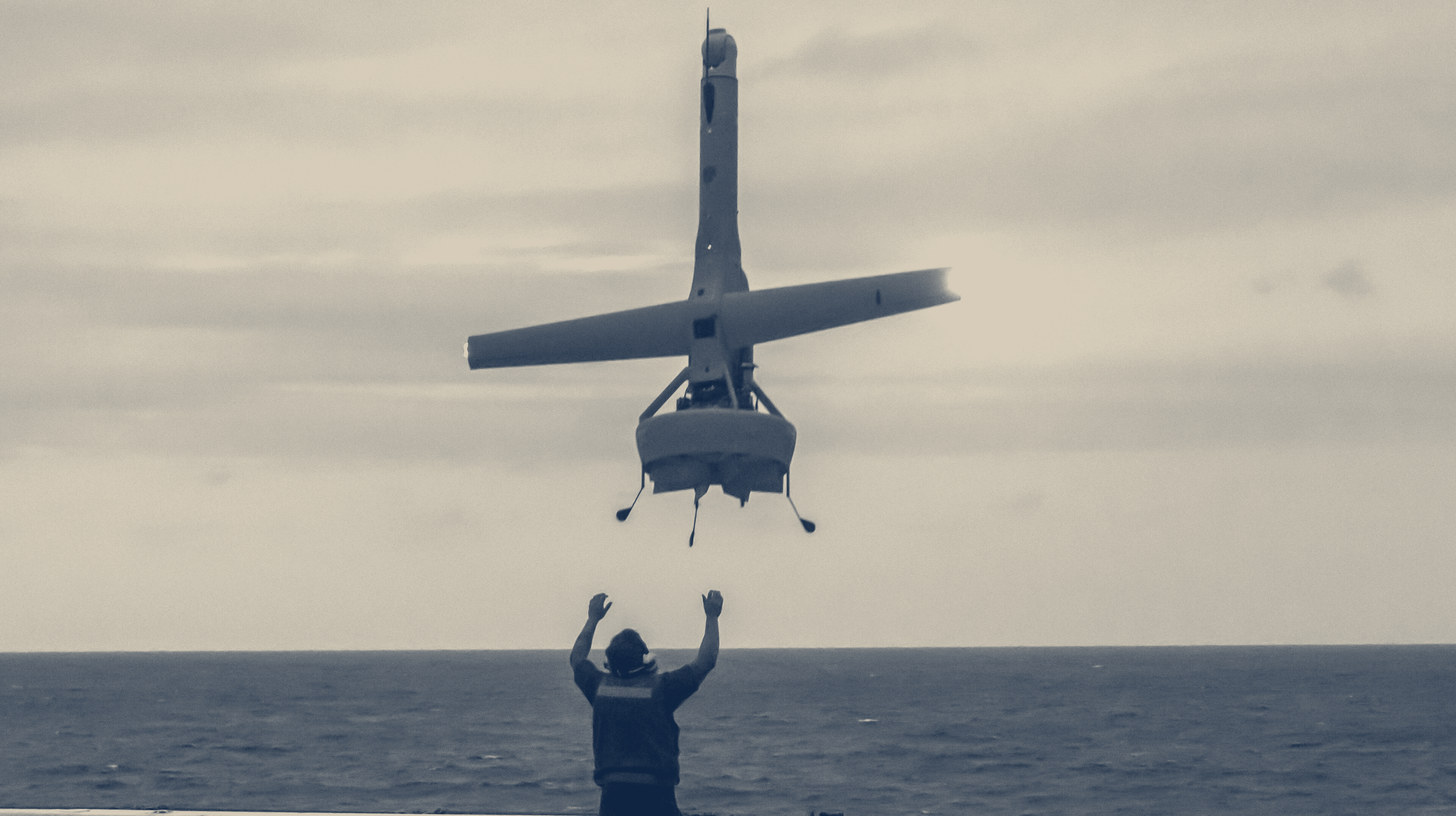 Two-tone U.S. Marine Corps photo of a V-BAT drone hovering down toward the deck of USS Green Bay as a sailor signals below.